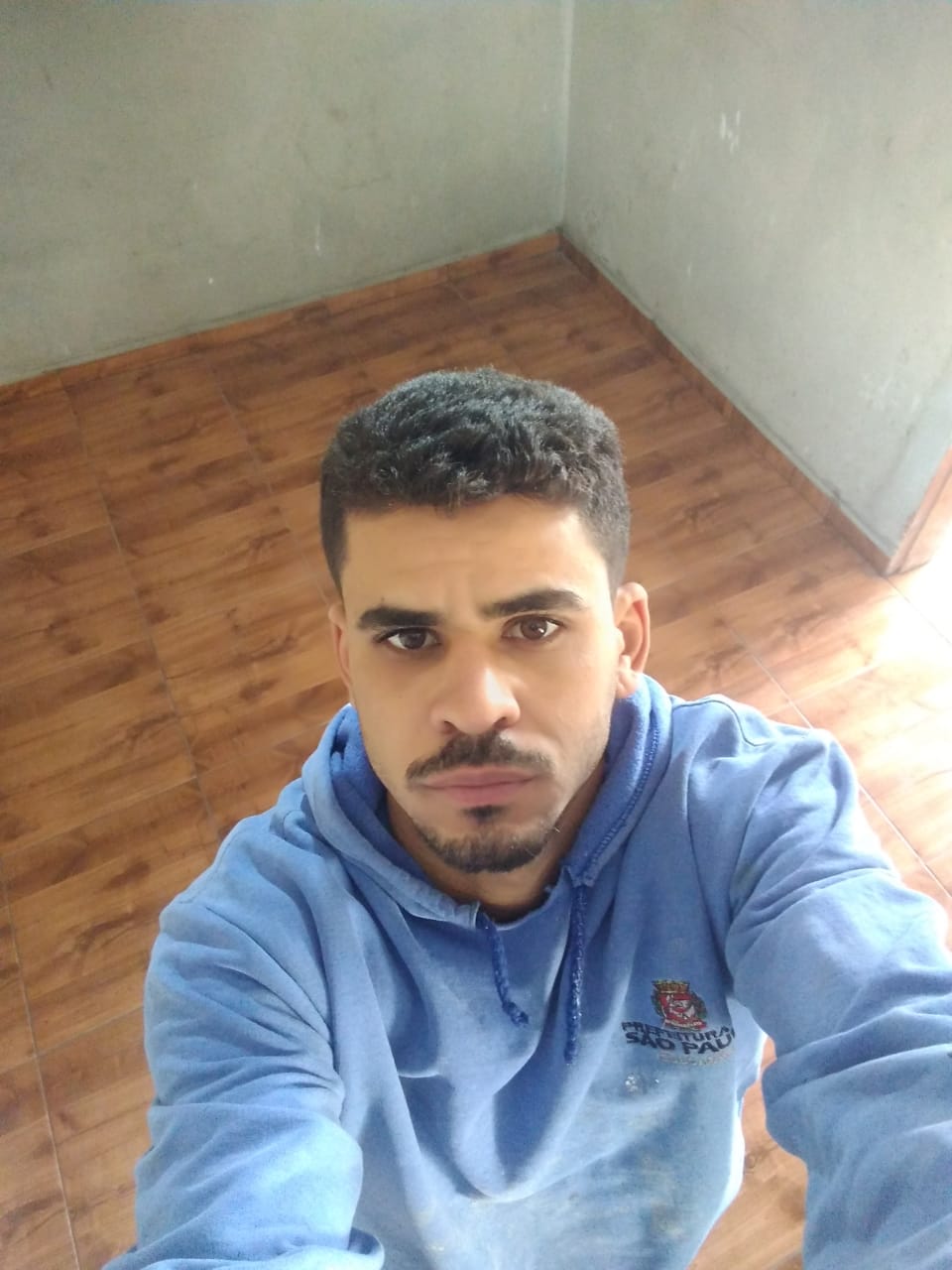 uma pessoa em um quarto com piso de madeira e parede em branco, usando um moletom azul com um logotipo no ombro