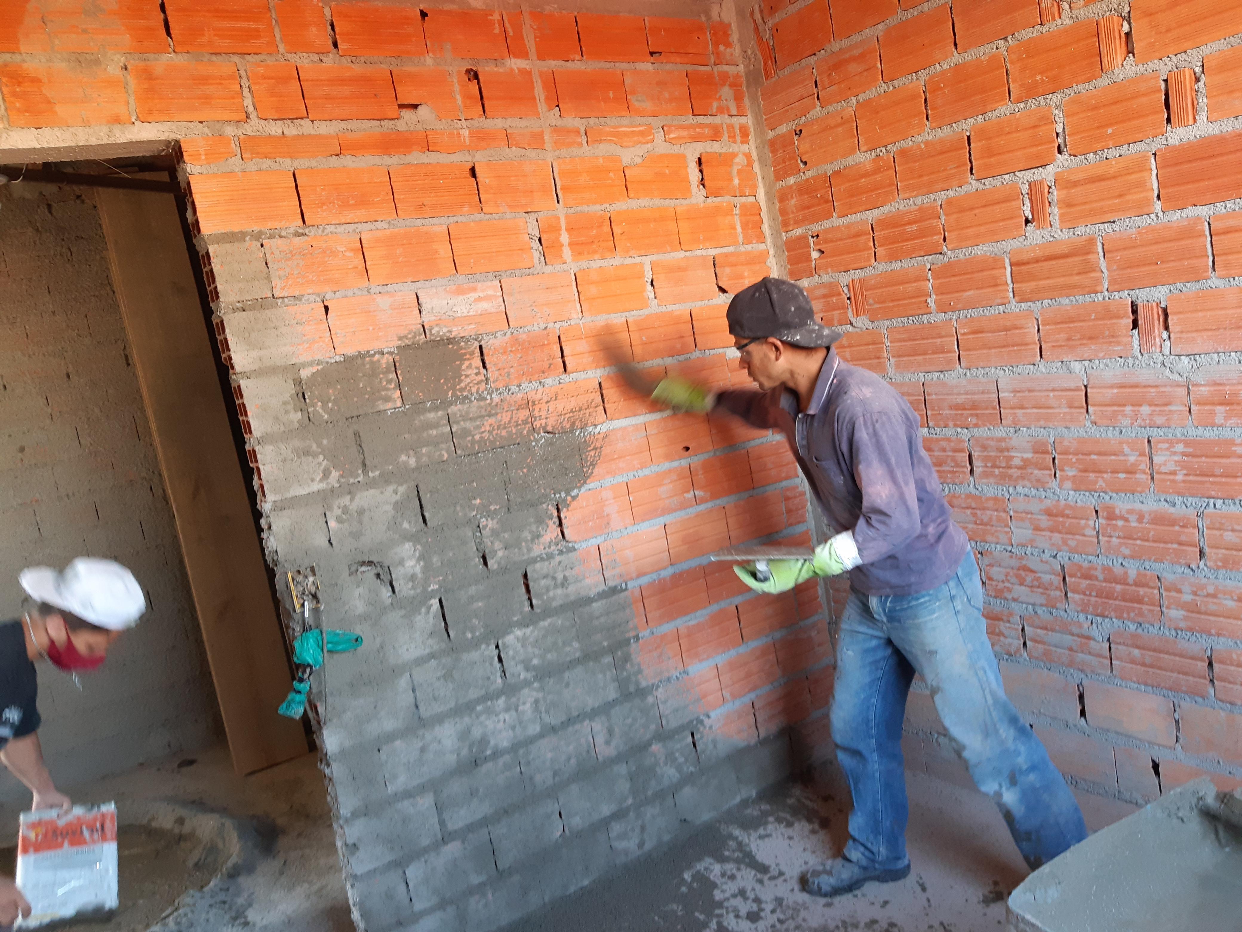 Trabalhadores em uma construção, um aplicando argamassa em uma parede de tijolos de barro, outro carregando um saco de material