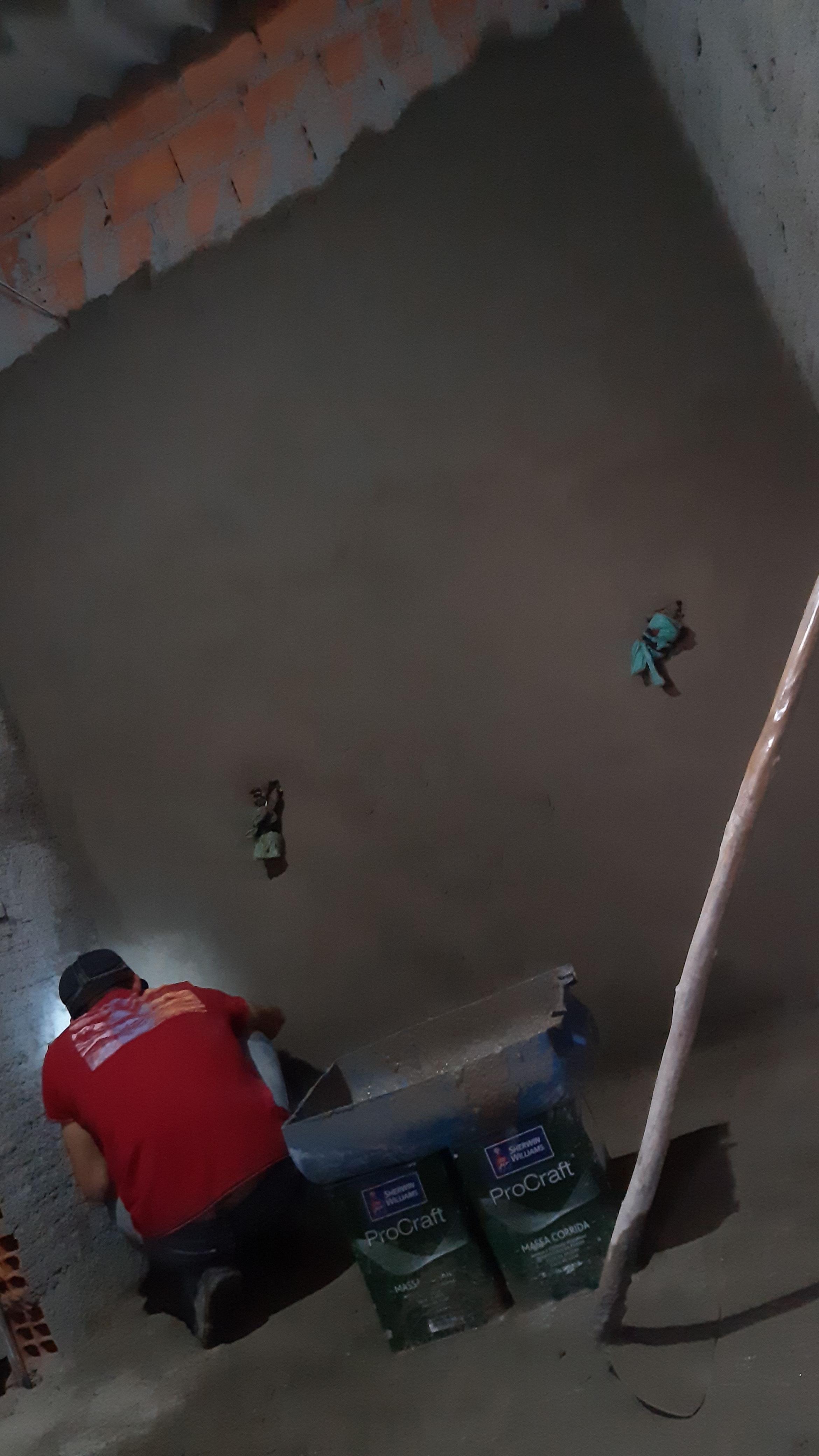 homem de camisa vermelha agachado aplicando massa em parede, com dois sacos de ProCraft Sher Williams no chão e tijolos aparentes na parte superior