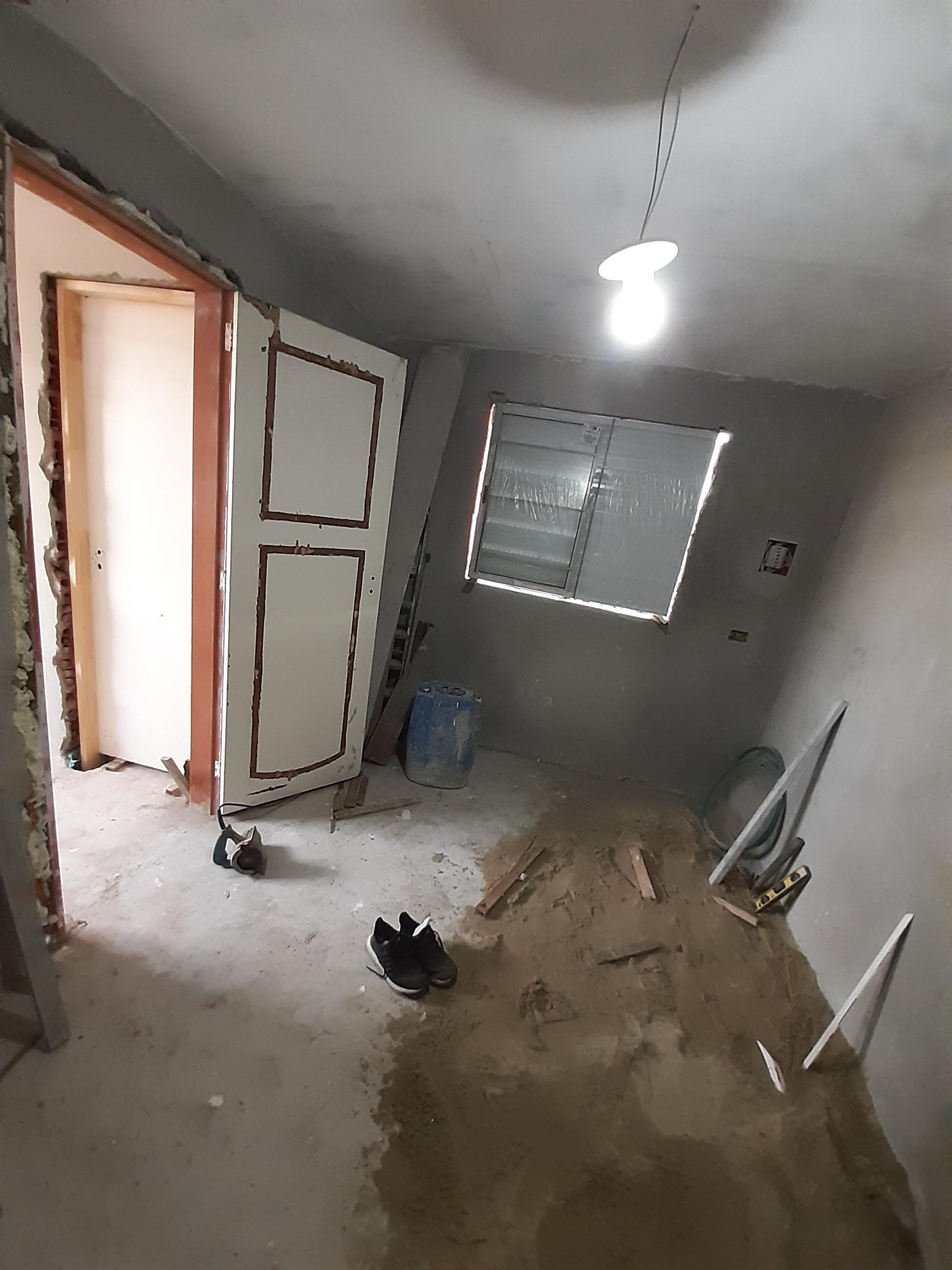 uma sala em construção com paredes cinzentas, uma porta aberta, uma janela, ferramentas e areia no chão