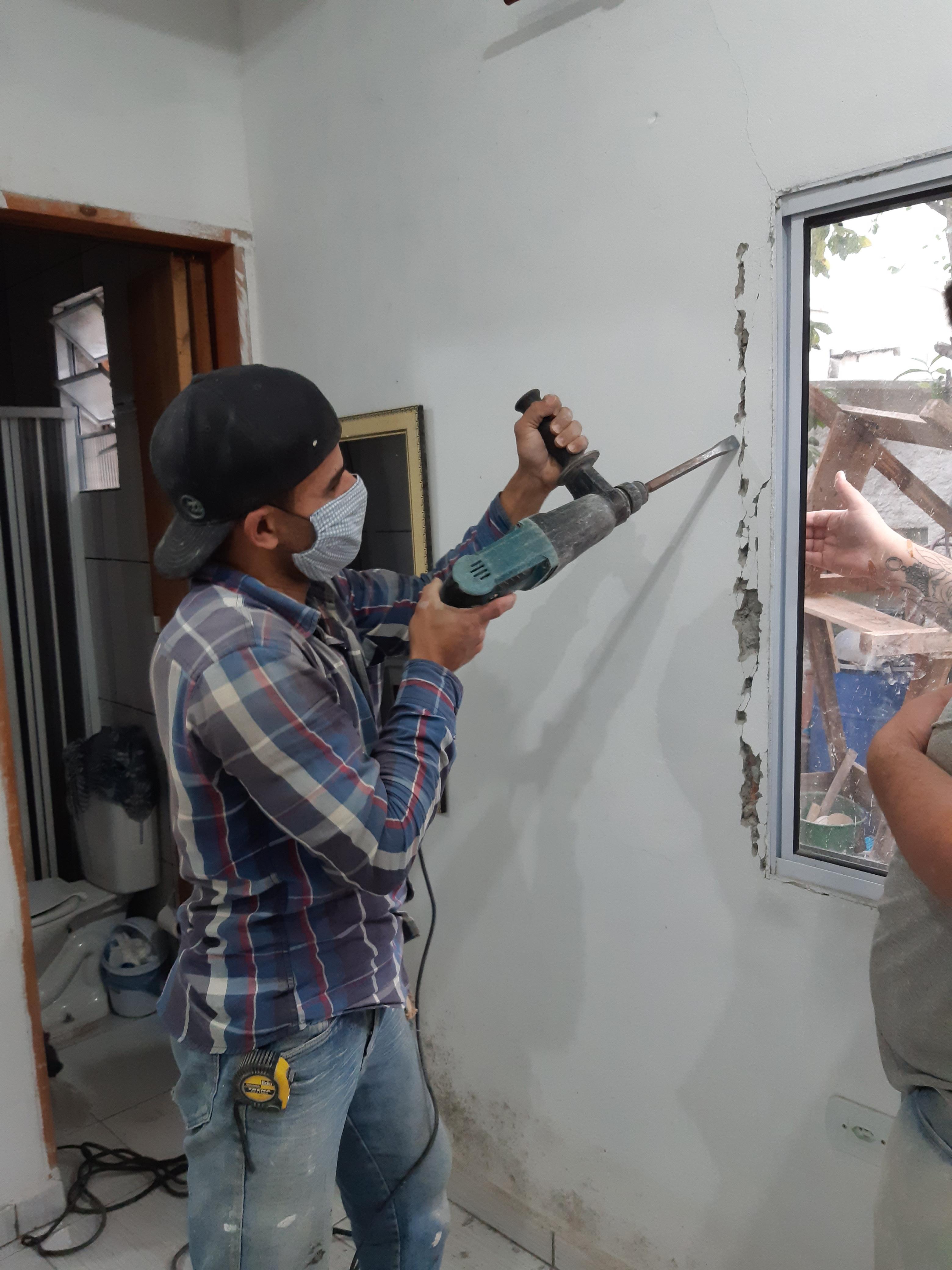 trabalhador usando furadeira na parede de uma construção, com outra pessoa assistindo e banheiro ao fundo