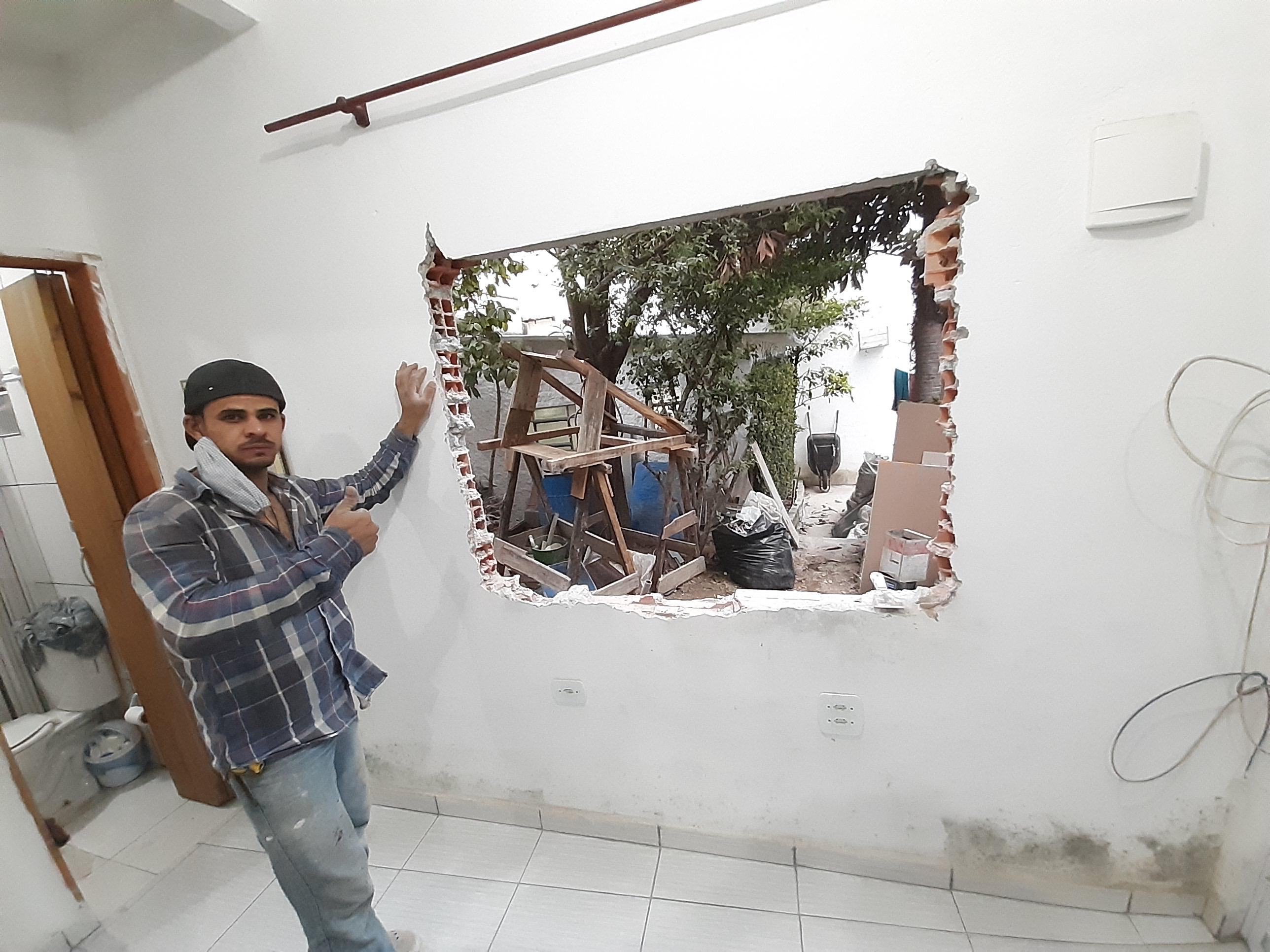 homem de boné e camisa xadrez em pé dentro de um cômodo com parede branca, apontando para uma abertura irregular feita na parede que dá para um quintal com madeiramento e árvores a