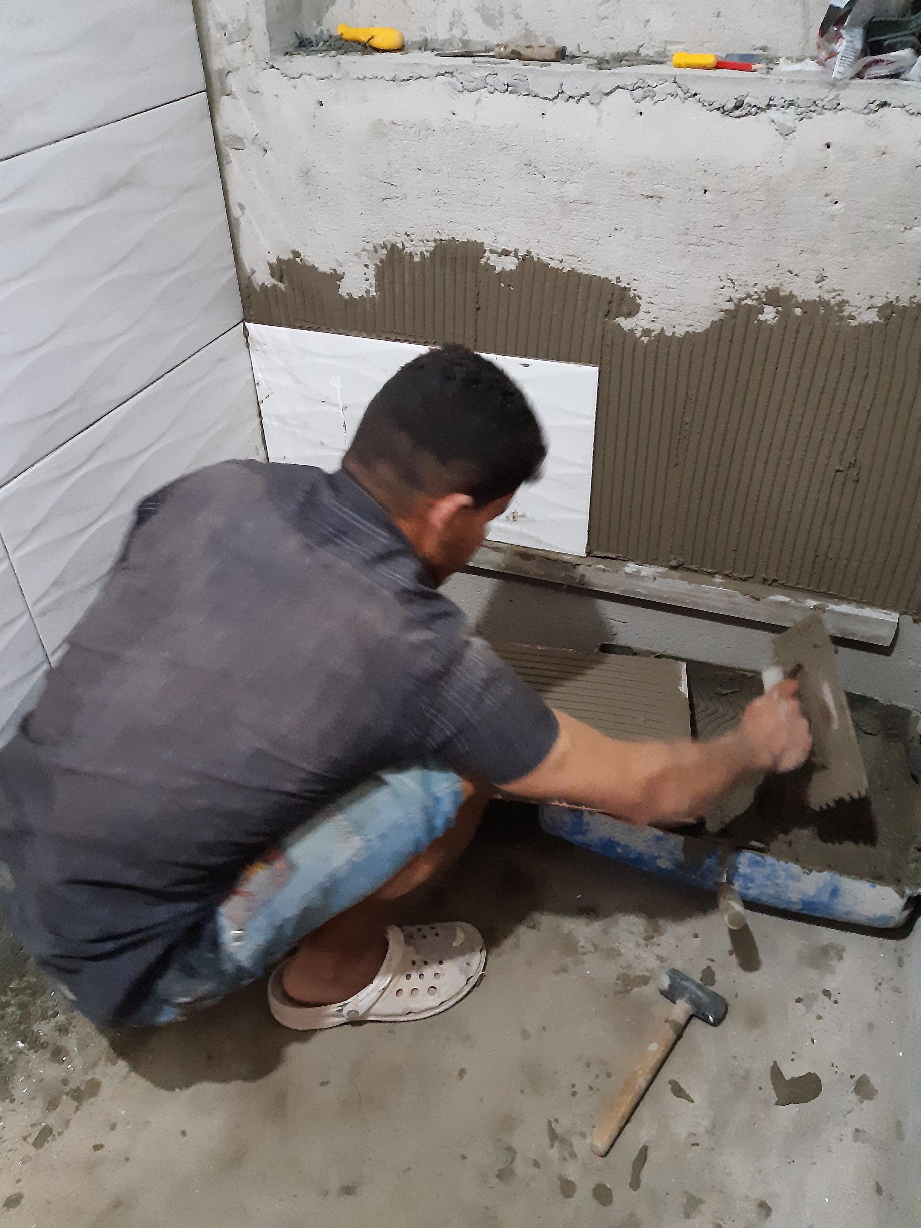 homem agachado aplicando argamassa em parede durante obra de revestimento cerâmico, com ferramentas no chão e azulejos brancos na lateral