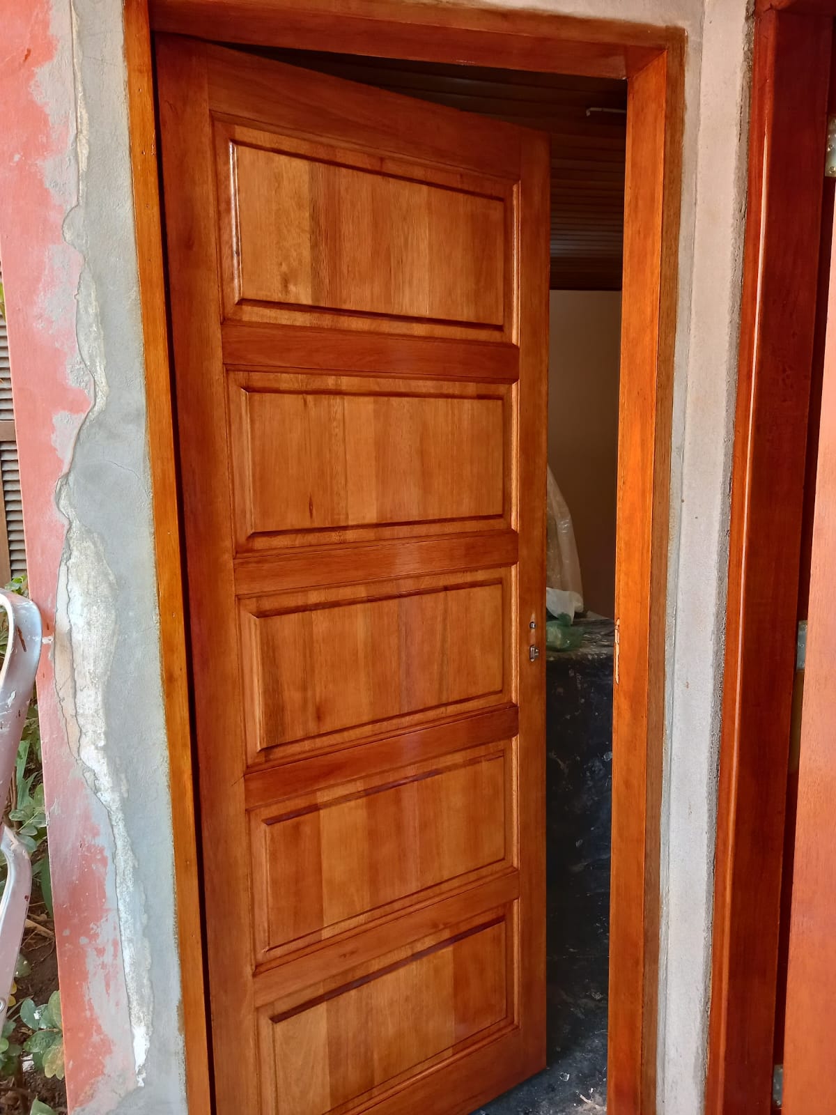 porta de madeira escura com quatro painéis instalada em batente de madeira, com parede de cimento cru ao lado