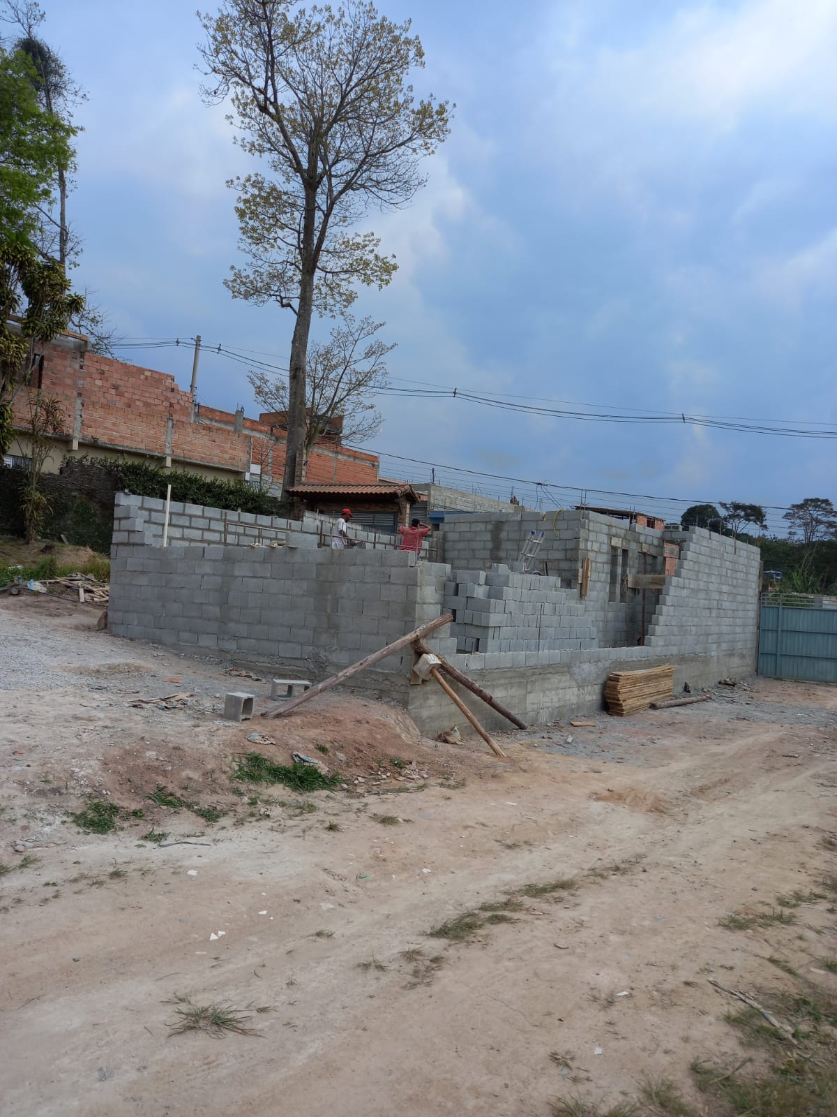 obra de construção civil com paredes de blocos de concreto, trabalhadores no topo e terreno de terra batida ao redor