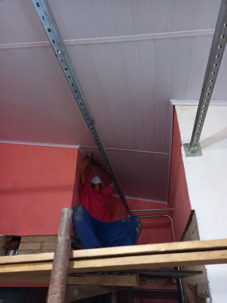 trabalhador em andaime instalando forro de PVC branco em ambiente com paredes vermelhas e perfis metálicos