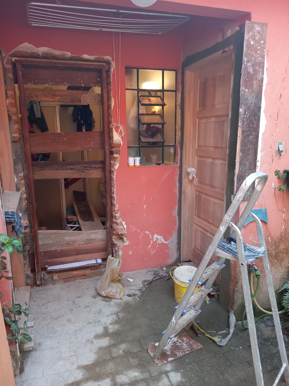 Imagem de uma área externa de uma casa em reformação, com portão de madeira desmontado, pintura em progresso na parede de cor rosa, uma porta de madeira, uma escada de alumínio, e 