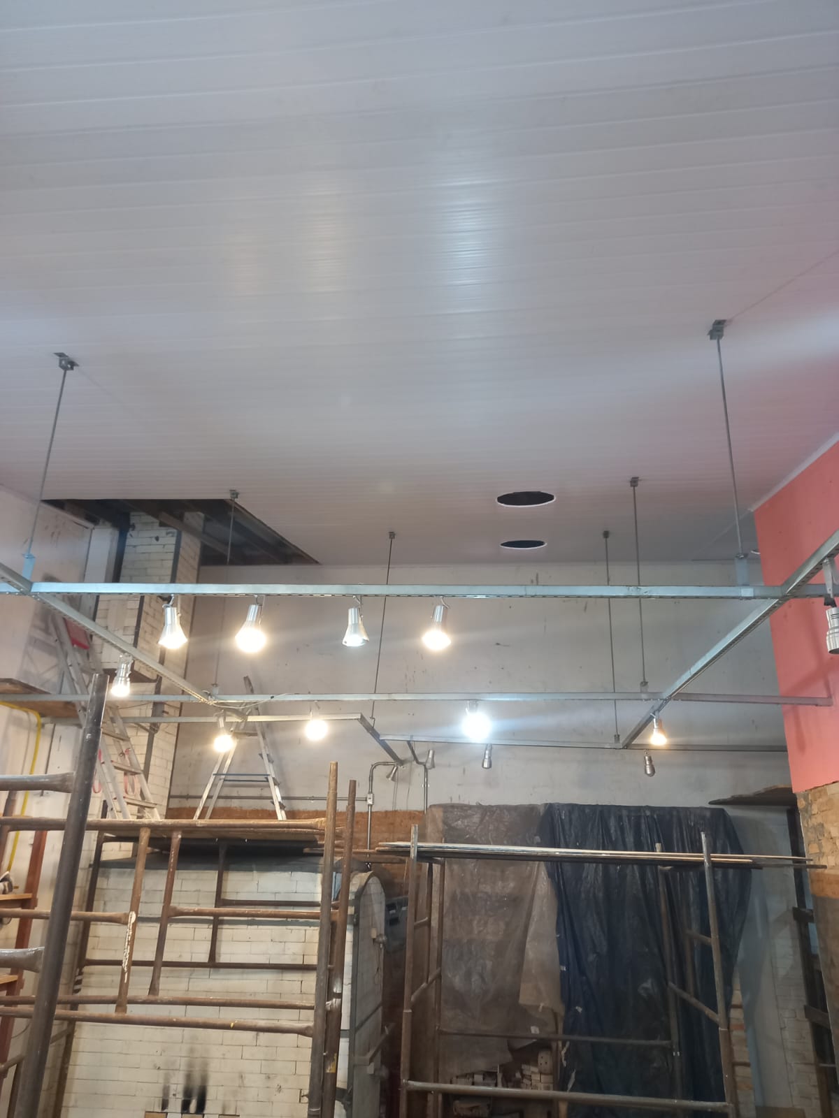 Interior de uma construção em andamento com telhado, lâmpadas, escadas de madeira e paredes brancas e de cor de laranja