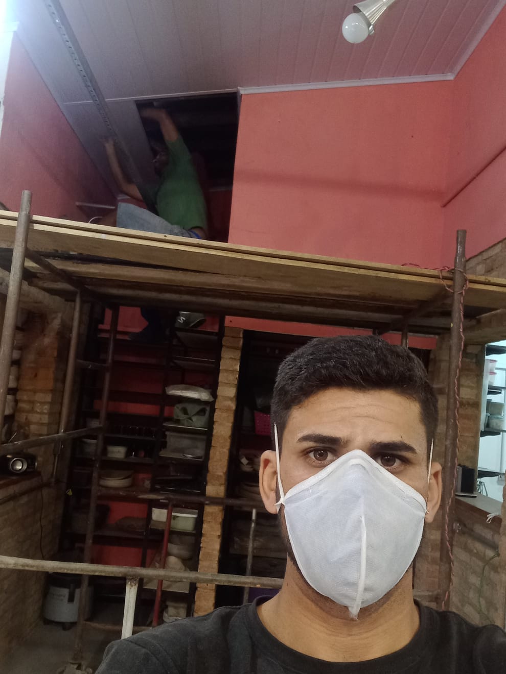uma pessoa com máscara no centro, trabalhadores em uma estrutura de madeira e uma parede vermelha no fundo