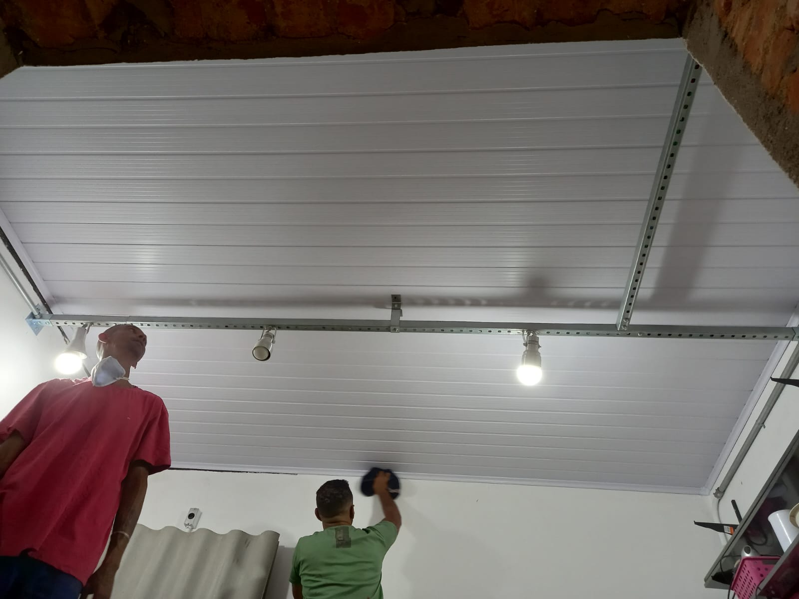 dois homens trabalhando na instalação de um forro de PVC branco em um ambiente com estrutura de metal aparente e iluminação