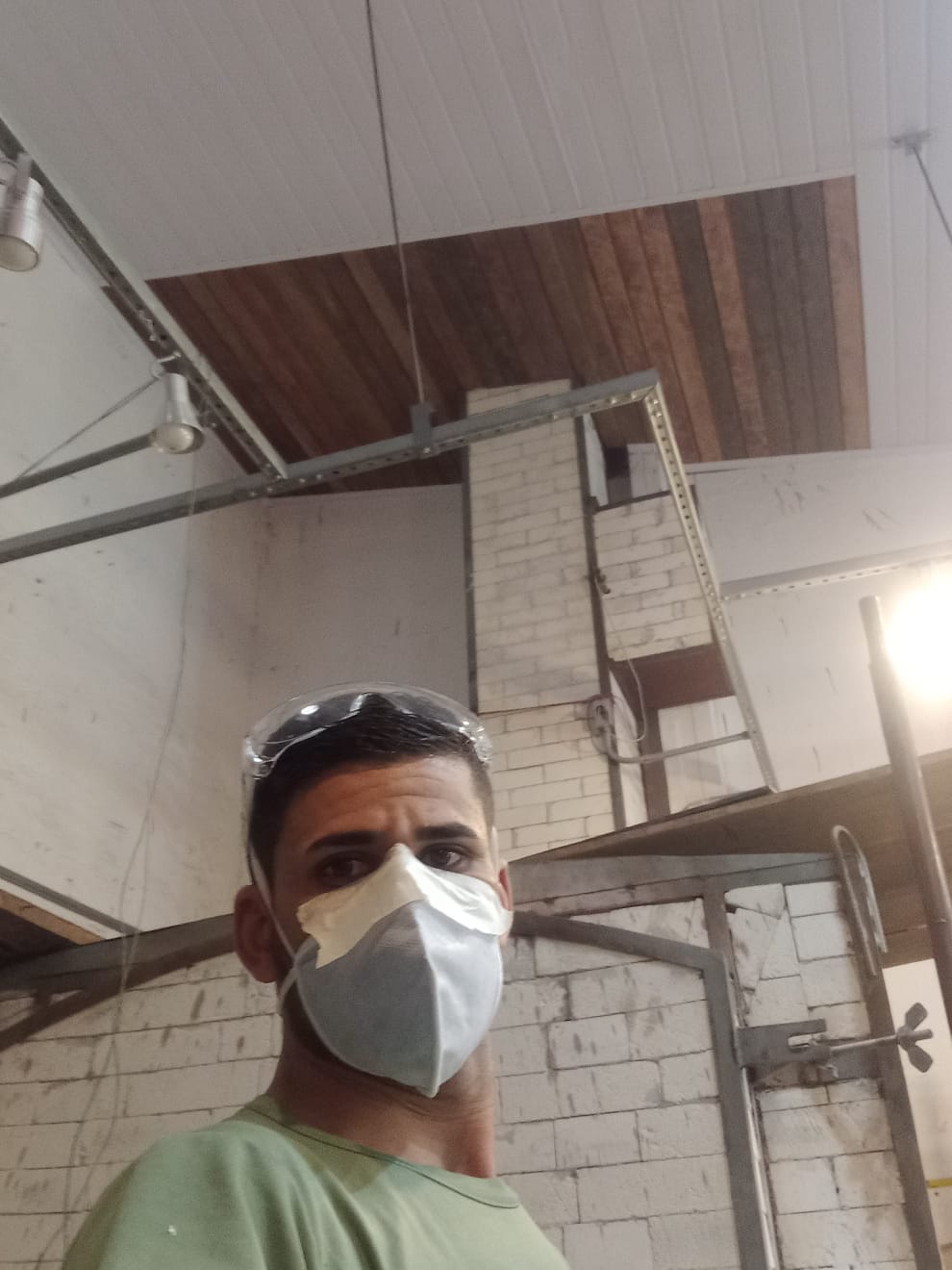 homem de camiseta verde usando máscara e óculos de proteção na cabeça, em ambiente interno com paredes de tijolos aparentes, estrutura metálica e forro de madeira