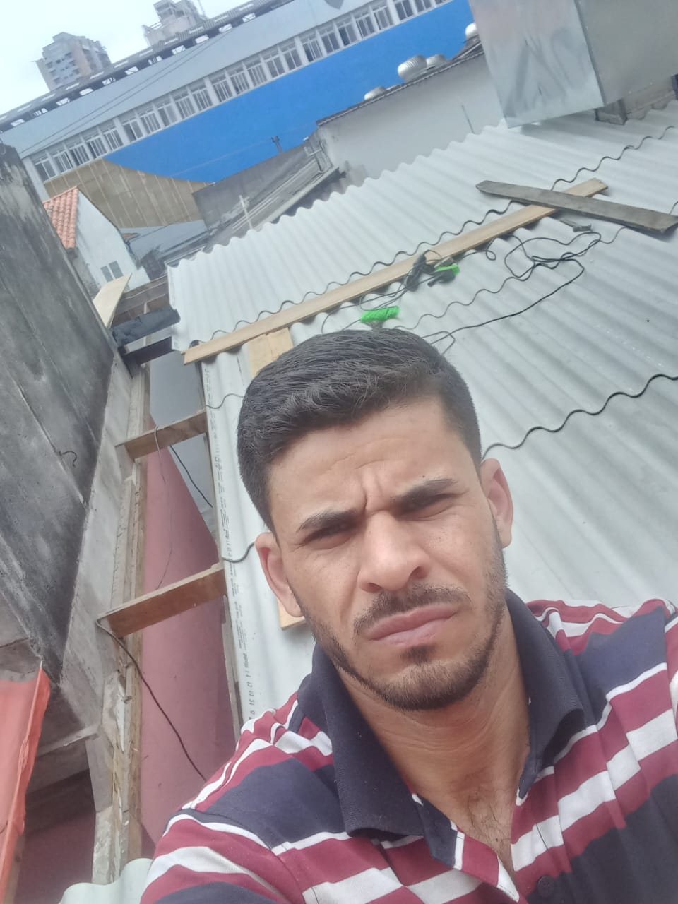 selfie de um homem com barba usando camisa listrada em frente a um telhado de telhas de fibrocimento e prédios ao fundo