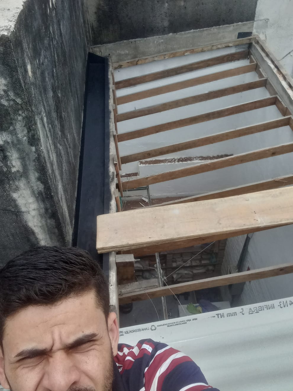 uma foto de um homem com barba e roupas listradas, perto de uma estrutura de madeira e concreto, com um painel de plástico com texto no fundo