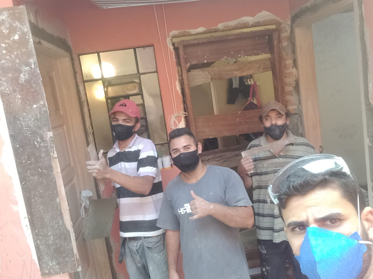 três trabalhadores com máscaras em uma construção em andamento, uma janela destruída e outra fechada, todos com os dedos em sinal de ok