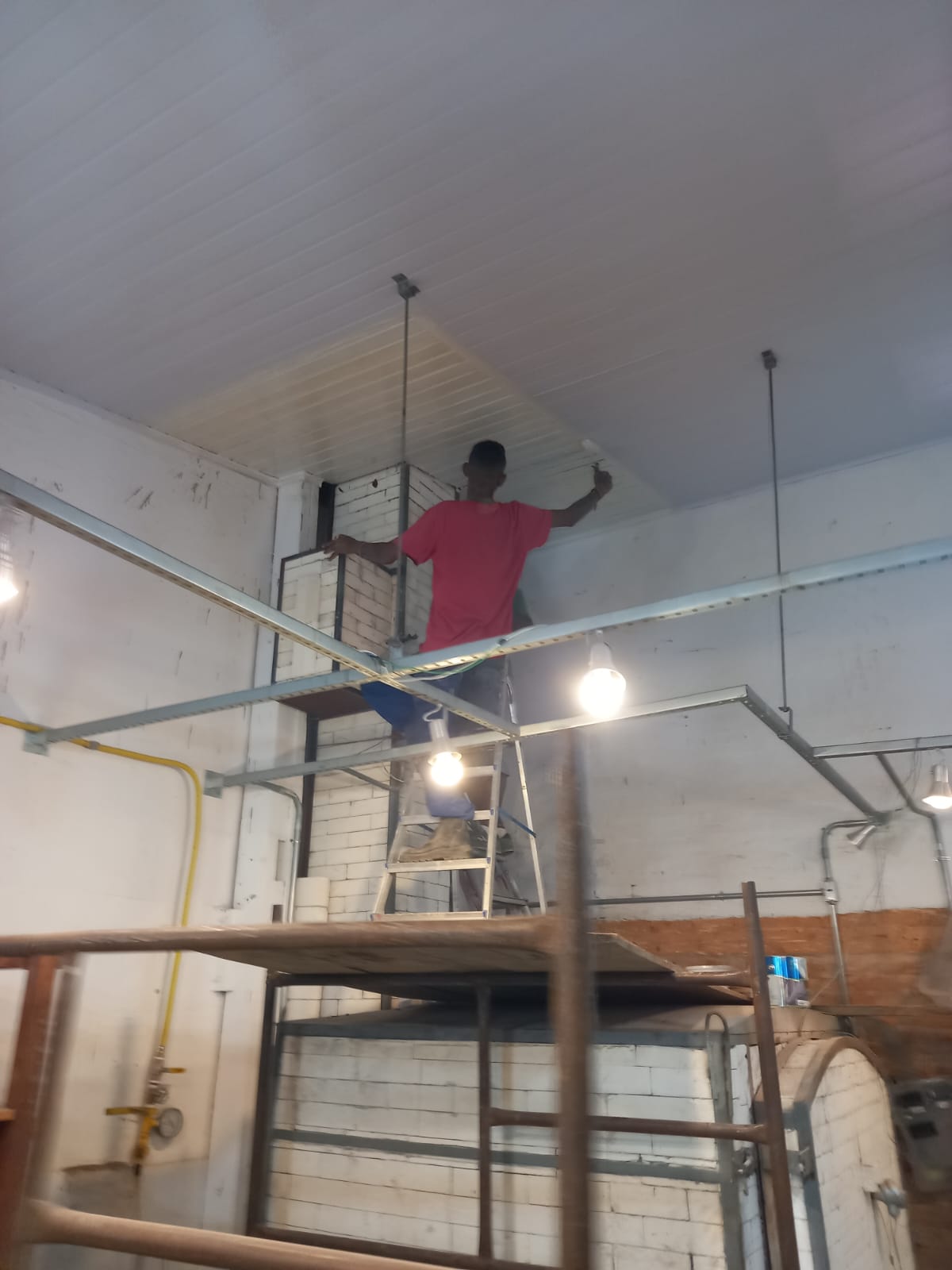 um trabalhador pintando o teto de uma construção industrial com escada e escada-rolante